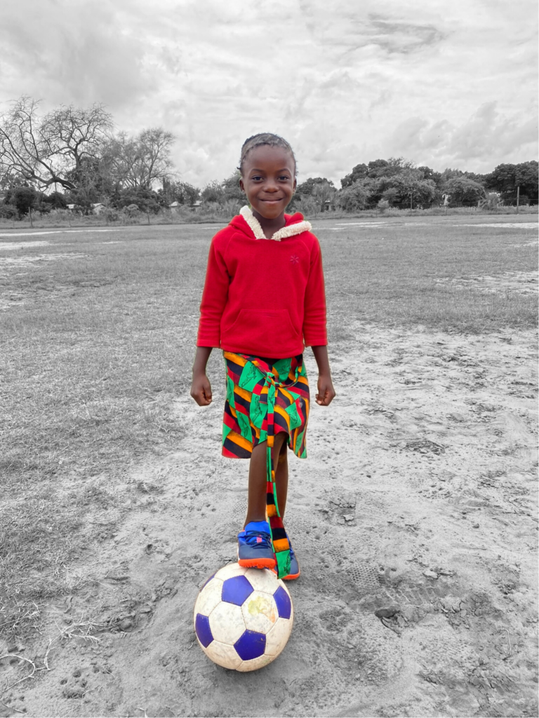 A young girl standing with a football at her feet.